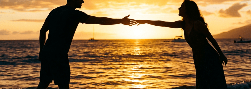 A man and woman reaching out their hands to each other on a beach at sunset.