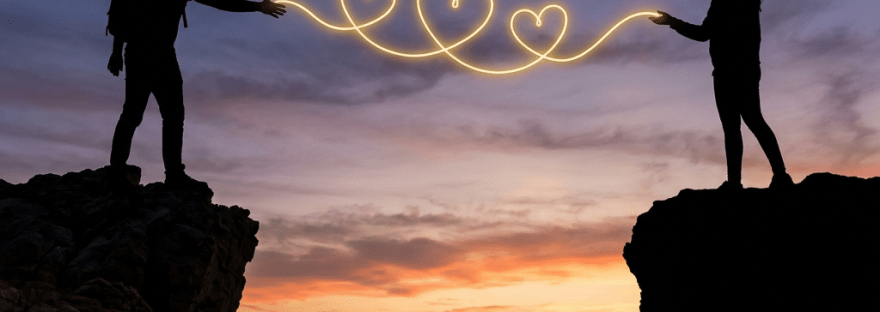 Silhouettes of a man and woman on cliff edges connected by glowing light hearts at sunset